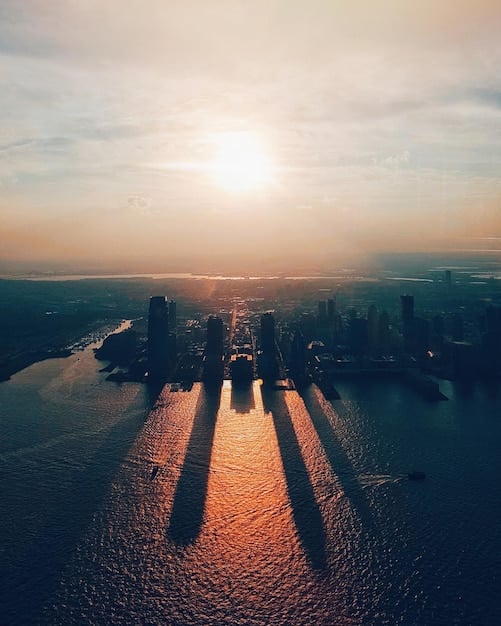 An aerial view of a sprawling metropolitan area at sunset, showing a mix of dense skyscrapers in the center gradually transitioning to residential suburban neighborhoods with parks and tree-lined streets on the outskirts.